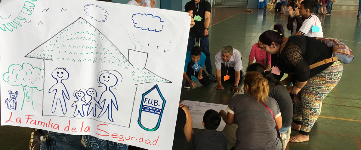 Familias desarrollando din�mica de grupo y un cartel en primer plano con una casa dibujada, dentro de esta una familia y bajo la misma el texto  `La familia de la seguridad`